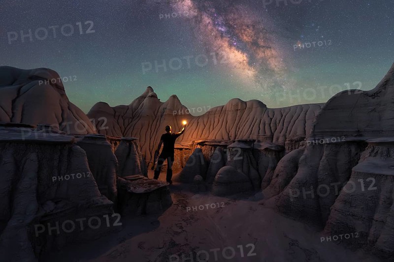 A lone figure holding a light stands amidst Goblin Valley's unique sandstone formations, beneath a stunning starry sky and the Milky Way, creating a mystical night scene