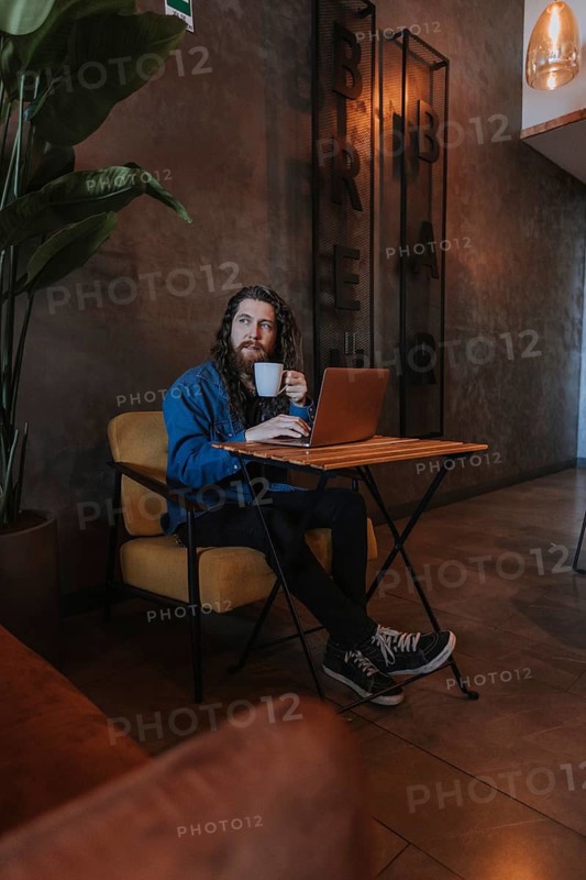 A man relaxes with a coffee in a cozy cafe, sitting with a laptop and looking contemplative. The ambiance is warm and inviting, perfect for work or leisure
