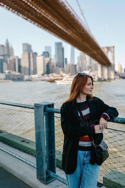 Woman in casual attire stands by a fence, enjoying the city skyline and bridge in New York. Her relaxed pose captures a mood of tranquility against the urban landscape