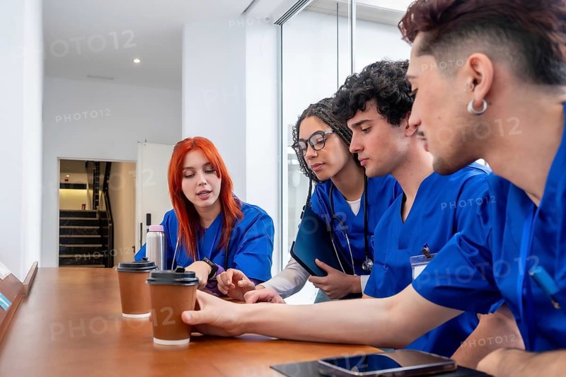 Group of young medical professionals and students in blue scrubs having an informal discussion and learning session during a coffee break, collaborating as a team in a modern hospital setting