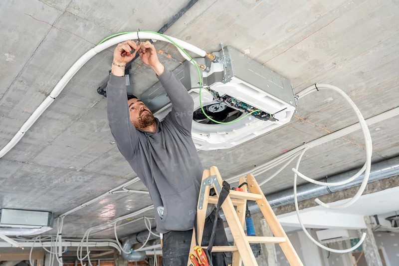 Professional hvac technician working on a modern air conditioning unit installation in a construction site, wiring and connecting pipes on a concrete ceiling of a new building