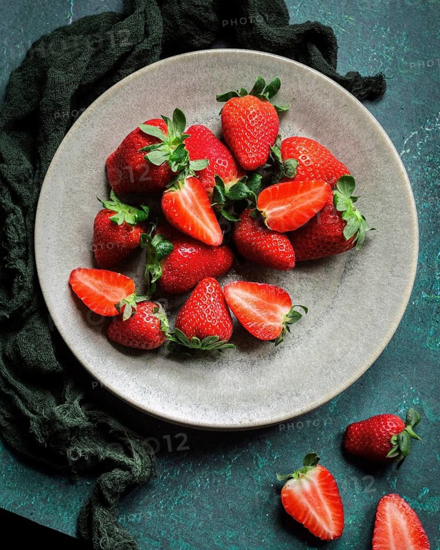 A ceramic plate filled with vibrant red strawberries, some cut in half, placed on a textured green surface with a dark green fabric accentuating the color of the fresh fruit