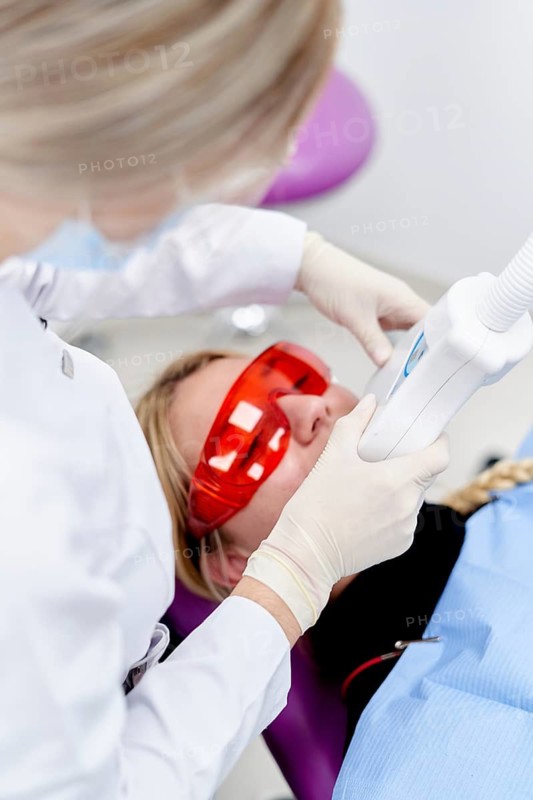 A dentist in a dental clinic uses a laser device for teeth whitening. The patient is wearing protective red glasses and a blue bib, highlighting a modern dental procedure