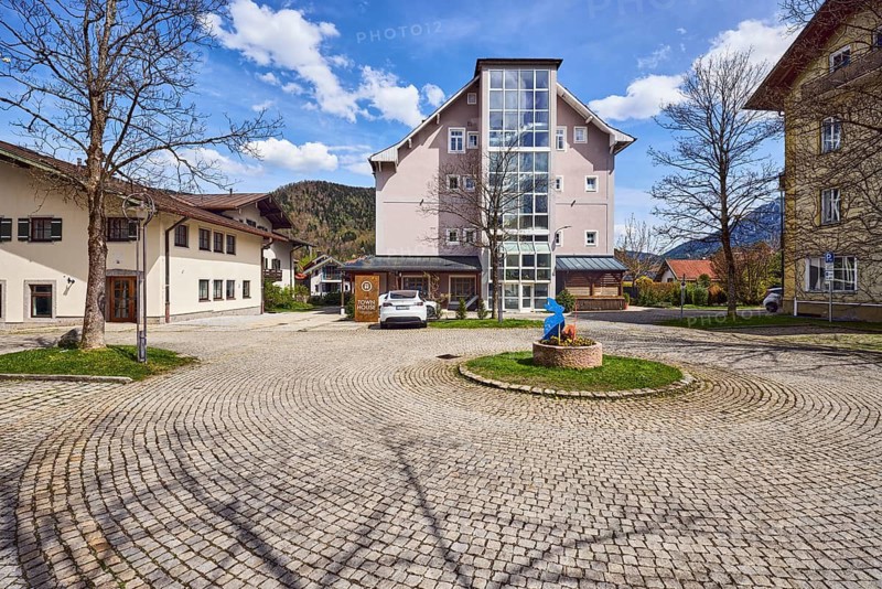 Hotel The Townhouse, commercial building, residential building, modern architecture, general development, parking, square, turning loop, car, paved roundel, flower pot, Easter decoration, street made of paving stones, bare winter trees, hilly landscape, coniferous forest, blue sky, cumulus clouds, town hall square, Ruhpolding, district of Traunstein, Bavaria, Germany