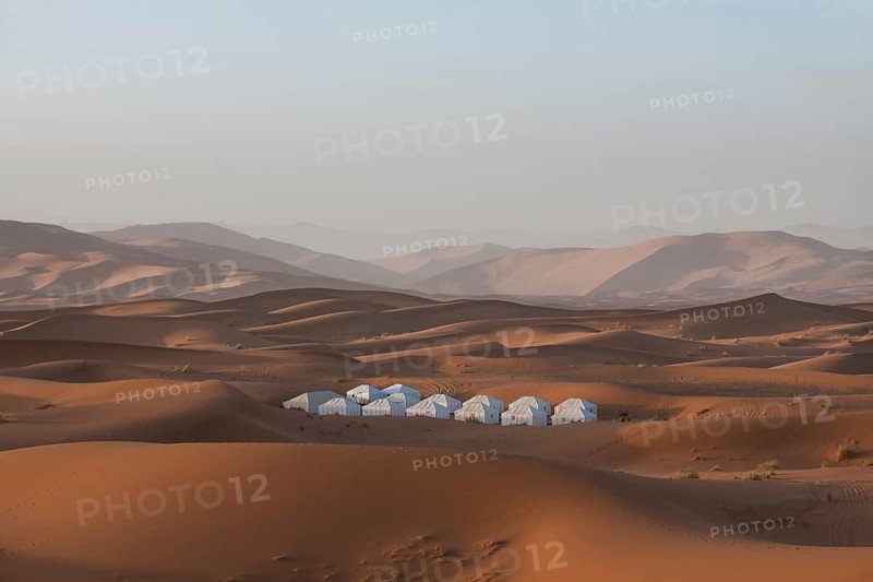 A remote Merzouga desert camp nestled among vast, rolling sand dunes under a clear blue sky. The landscape is serene, showcasing the beauty and tranquility of the desert environment in Morocco