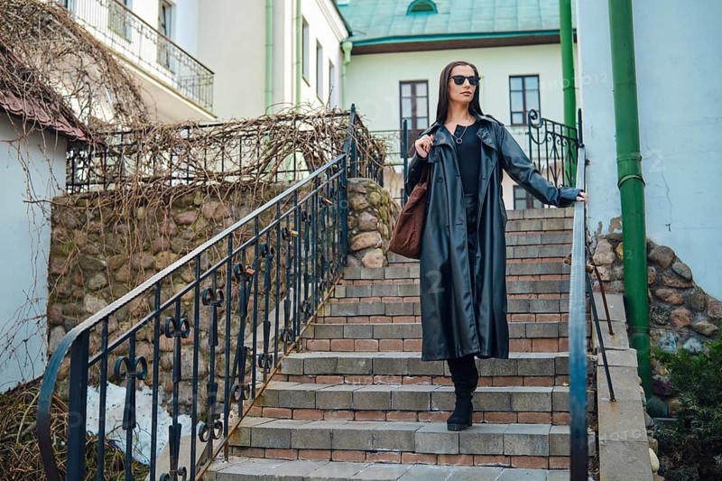 A woman is going down a set of stairs in a city environment. She wears sunglasses and holds a brown bag. The stairs are bordered by a railing. Buildings are in the background