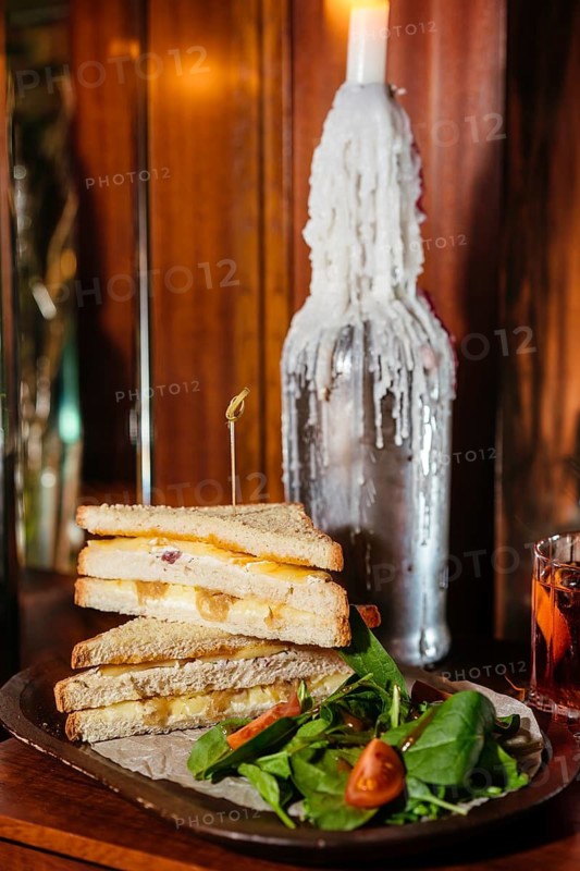 A deliciously stacked club sandwich filled with melted cheese and caramelized onions, served with a fresh side salad. A rustic candle-lit setting and a drink create a cozy, vintage ambiance