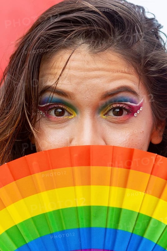 Close-up of a woman with vibrant rainbow makeup and jewels, holding a rainbow fan, symbolizing LGBTQIA+ pride and diversity. The image exudes celebration and joy