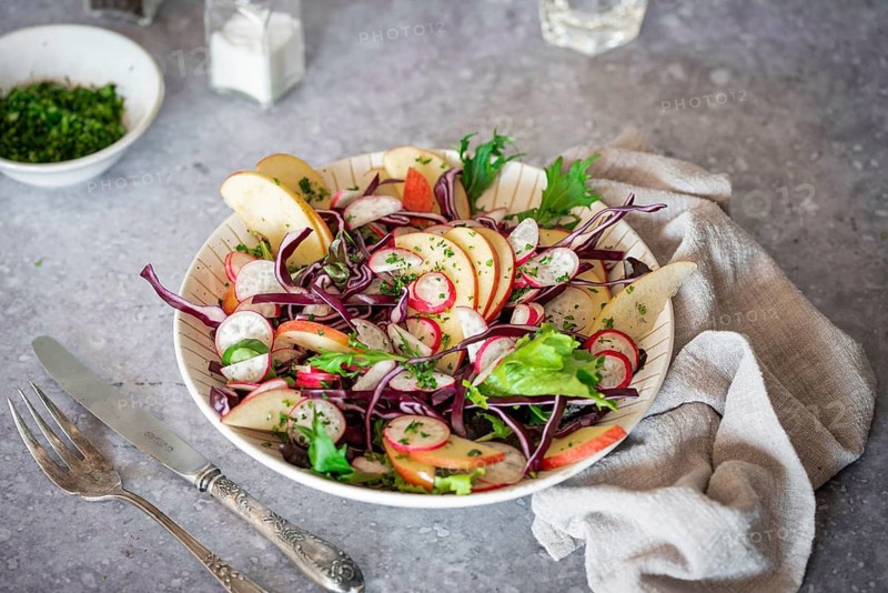 A vibrant salad featuring red cabbage, radishes, apple slices, and mixed greens. Served on a rustic table with a vintage cutlery set, creating a fresh and inviting look