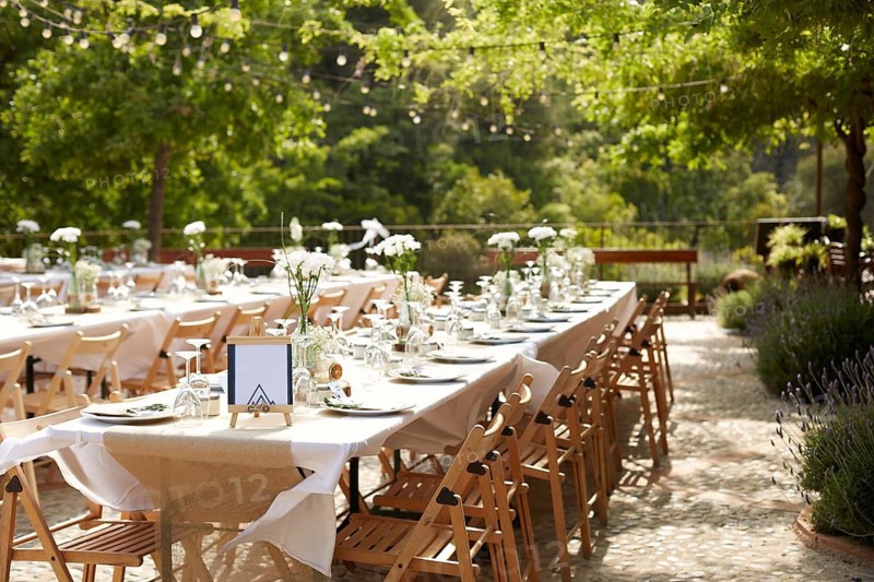 Elegant outdoor wedding setting featuring long tables adorned with white floral arrangements, rustic wooden chairs, and soft string lights under lush greenery