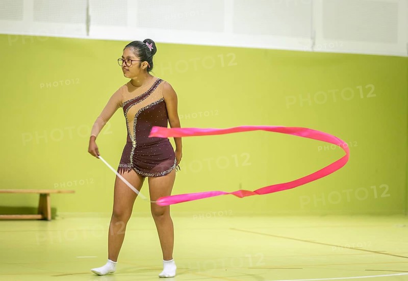 A disabled rhythmic gymnast performs a routine using a vivid pink ribbon, showcasing grace and precision in a green lit gymnasium, wearing a sparkling brown leotard