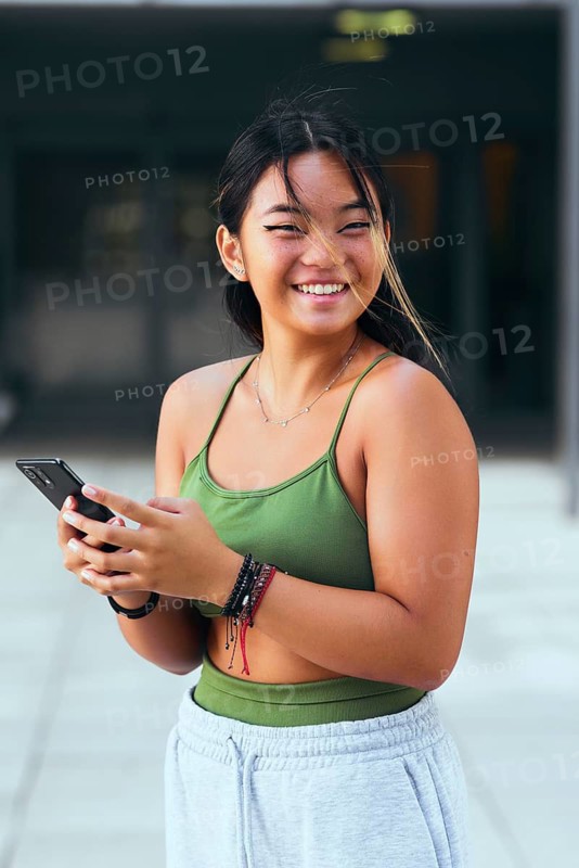 A cheerful teenager wearing a green top and gray sweatpants smiles while holding a phone. Strands of hair gently move in the breeze, capturing a lively urban moment