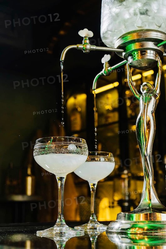 A vintage absinthe fountain dripping cold water into glasses, casting elegant reflections under warm bar lights. Ideal for cocktail or bar-related themes