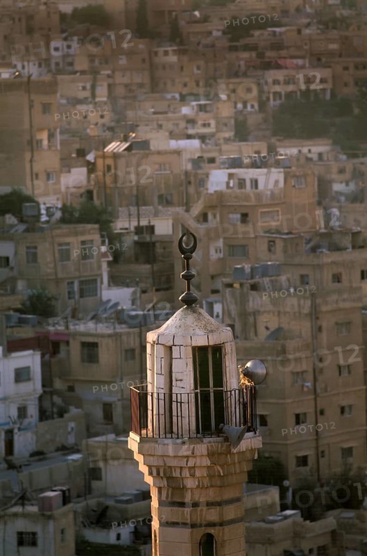 A mosque minaret with dawn sunlight set against a backdrop of traditional hillside houses in the Jordanian capital city of Amman, Jordan