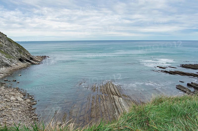 A serene coastal landscape showcasing a calm sea, striated rock formations, and lush green grass under a partly cloudy sky, depicting the untouched beauty of nature