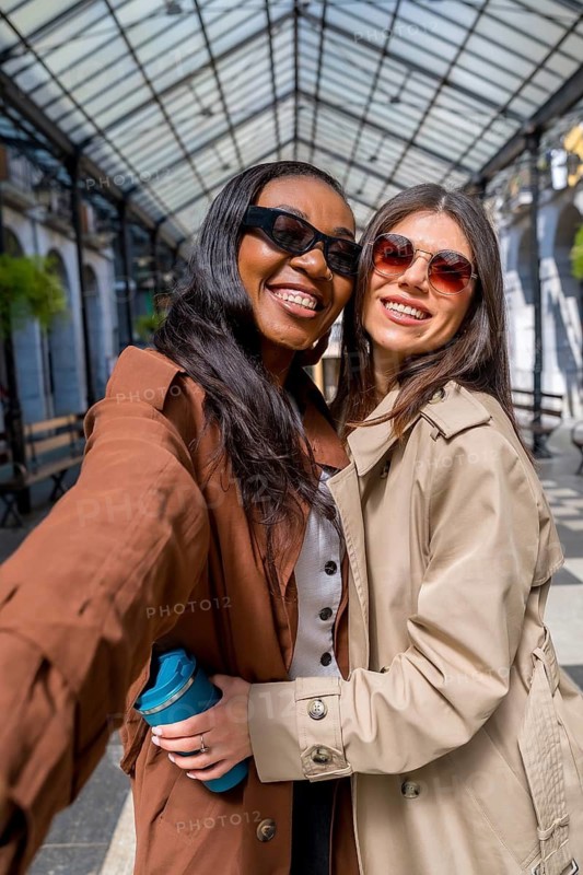 Two cheerful businesswomen wearing sunglasses are taking a selfie under a glass roof in a gallery