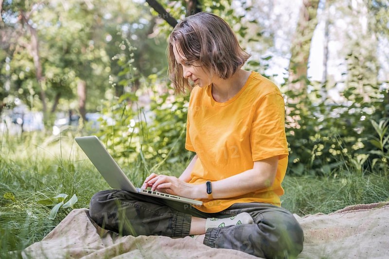Woman sits cross-legged on a blanket in a lush park, using her laptop. Surrounded by greenery, she enjoys a peaceful outdoor working session under the sun