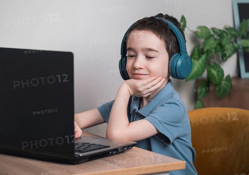 A young boy, smiling in headphones, engaged with a laptop. He is sitting at a wooden desk, surrounded by plants, enjoying an online learning experience in a cozy setting