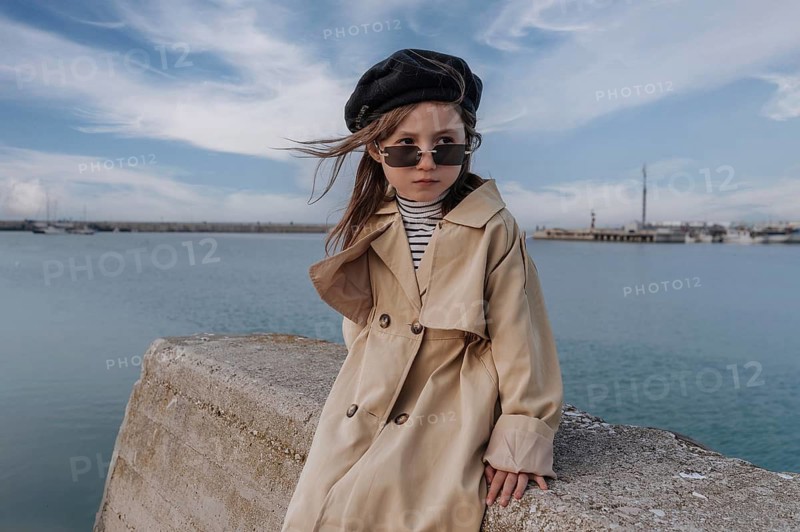 Young girl in a trench coat and beret poses stylishly by the sea in Civitanova, Italy. Her sunglasses add a fashionable touch, and the sky is beautifully clear