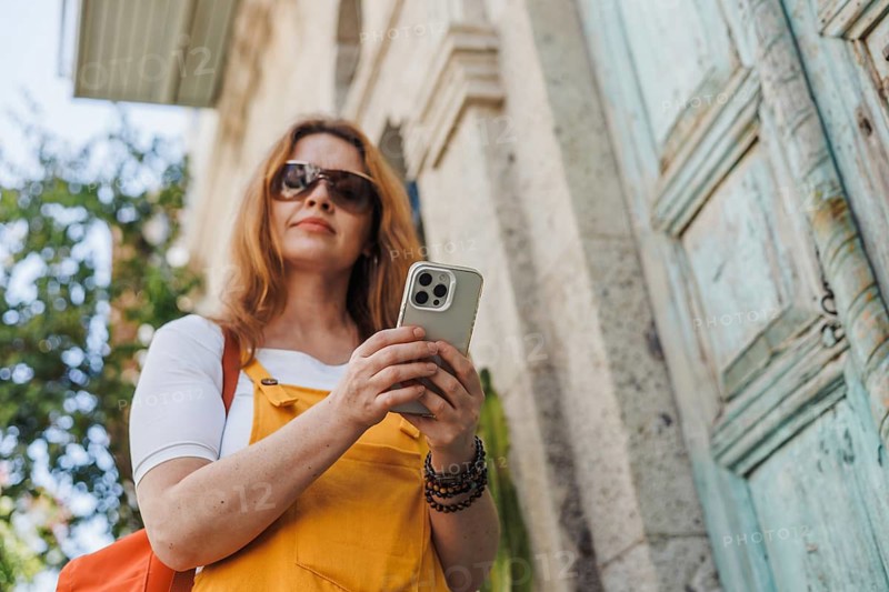 From below a woman in her 40s, dressed in orange and yellow, uses her smartphone as she explores the historic old city of Cesme, Turkey during her vacation