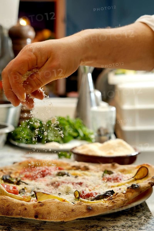 Cropped unrecognizable person sprinkling cheese over a freshly baked pizza in a home kitchen, with a focus on the food and hands, showing a glimpse of Italian culinary traditions