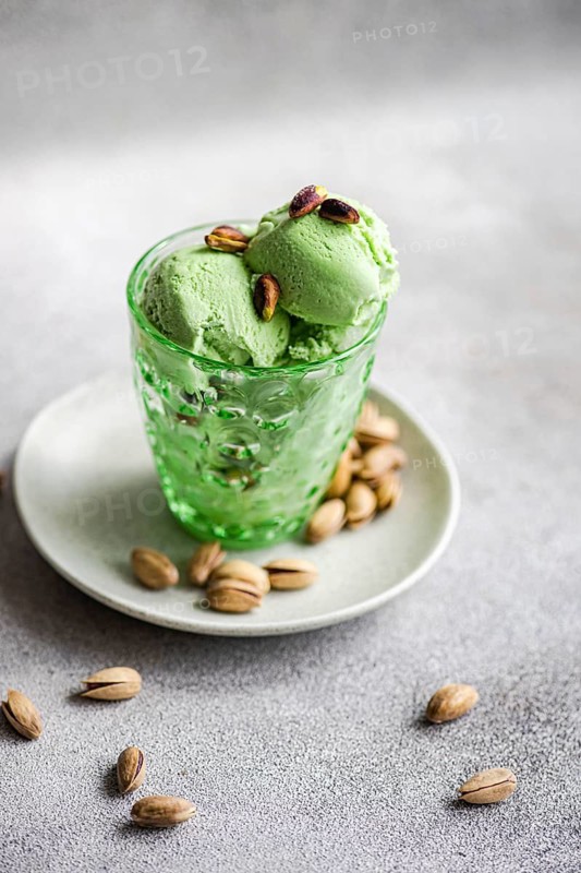A scoop of creamy pistachio ice cream in a textured green glass sits on a plate, surrounded by whole pistachios. Perfect for dessert and gourmet presentations