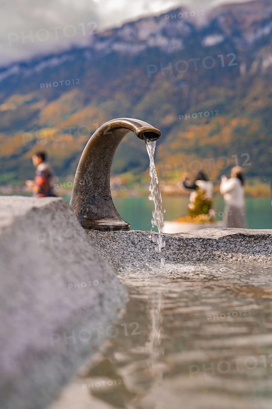 Fountain with running water, people at the lake with mountains in the background, Lake Brienz, Switzerland