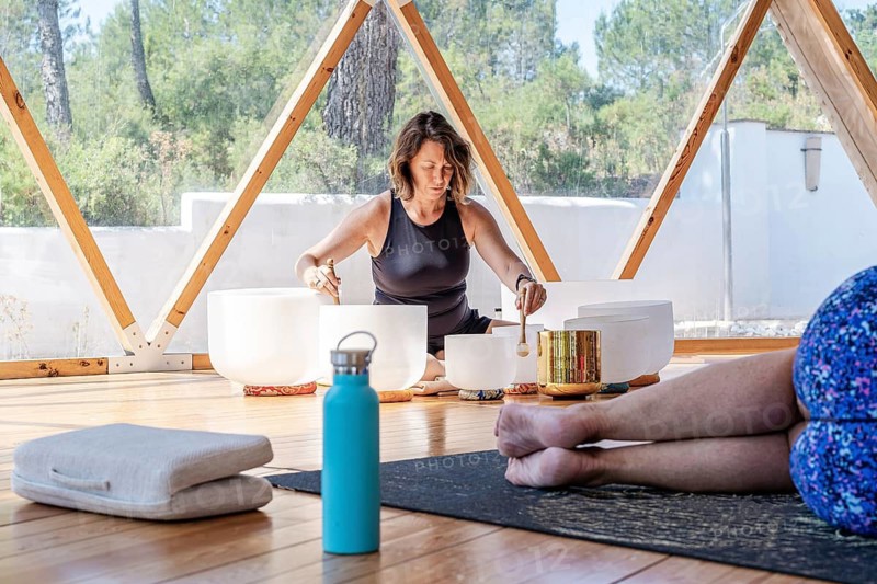 A woman sits cross-legged playing crystal singing bowls with mallets, creating soothing sounds in a serene setting. The scene promotes relaxation and holistic wellness