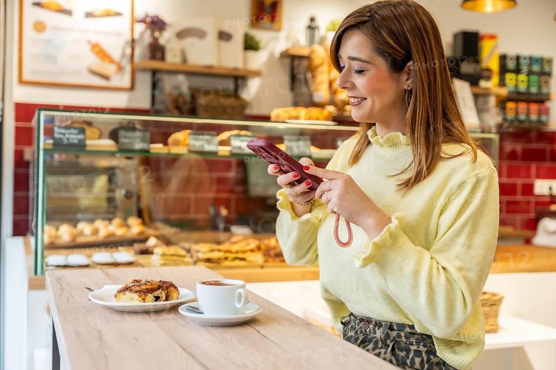 Smiling woman browsing her smartphone while relaxing at a bakery counter, enjoying a warm cup of coffee and a delicious cinnamon bun, showcasing leisure and modern lifestyle