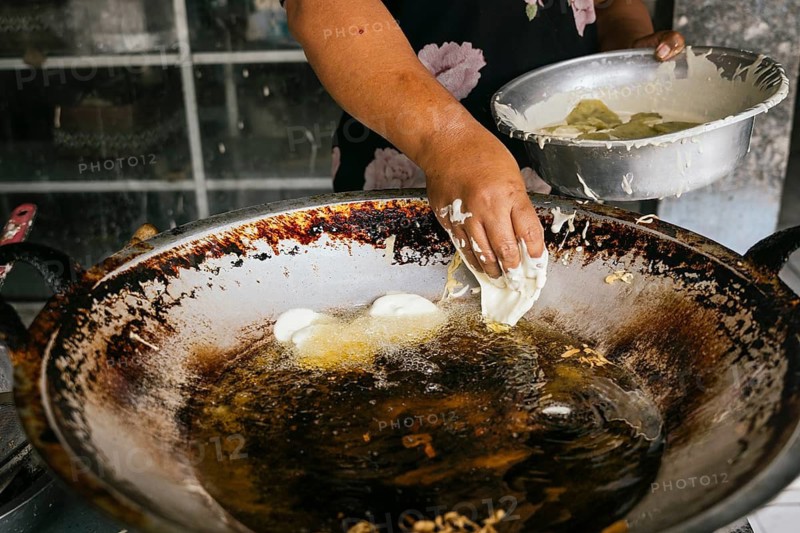 A street vendor expertly fries dough in a large, well used pan filled with hot oil. The scene captures the essence of traditional street food preparation of Bali