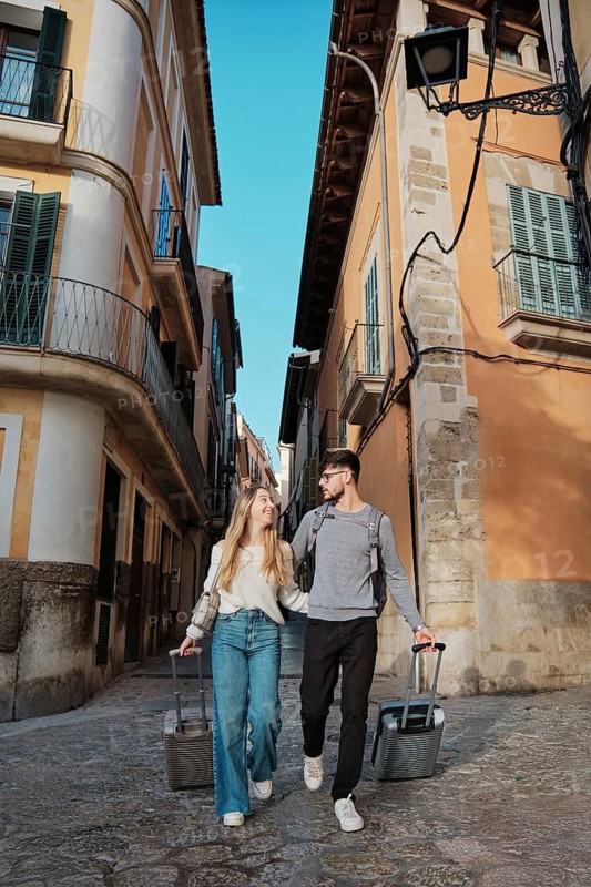 A young couple wanders through charming streets, searching for their perfect vacation home in Majorca, Spain. They are joyfully exploring a sunny Mediterranean setting