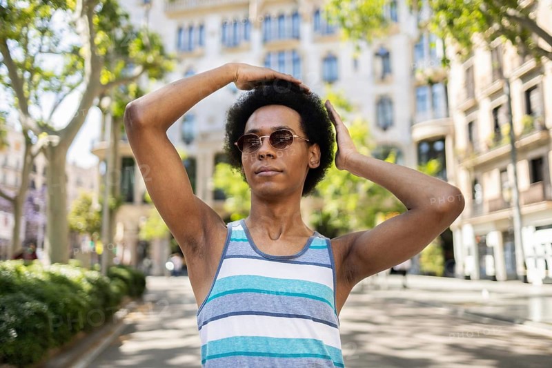 A confident non-binary person wearing sunglasses poses outdoors, enjoying a sunny day in the city. They are wearing a striped tank top, surrounded by urban architecture