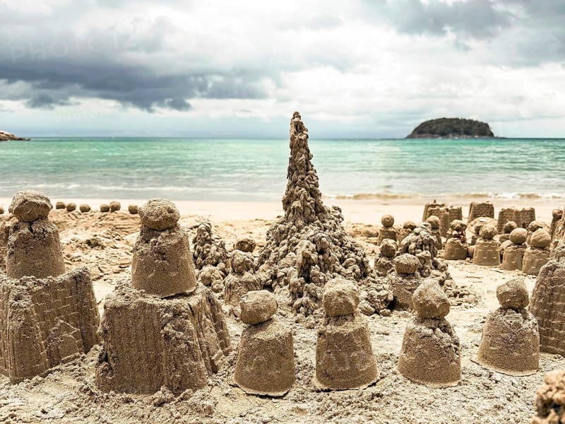 A collection of intricately crafted sand castles adorns the sunlit seashore. The ocean's gentle waves approach under a cloudy sky, creating a serene coastal backdrop
