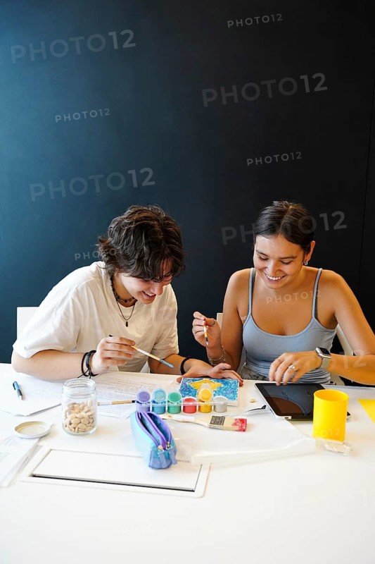 Two cheerful teenagers, a non-binary individual and a female, engage in a creative painting activity in a classroom. They are focused, sharing a moment of artistic expression and companionship with digital tablet