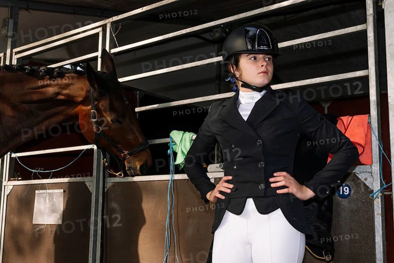 A teenage girl in traditional dressage attire stands confidently beside a dark horse. She wears a black riding jacket, white breeches, and a helmet, looking away from the camera with focus
