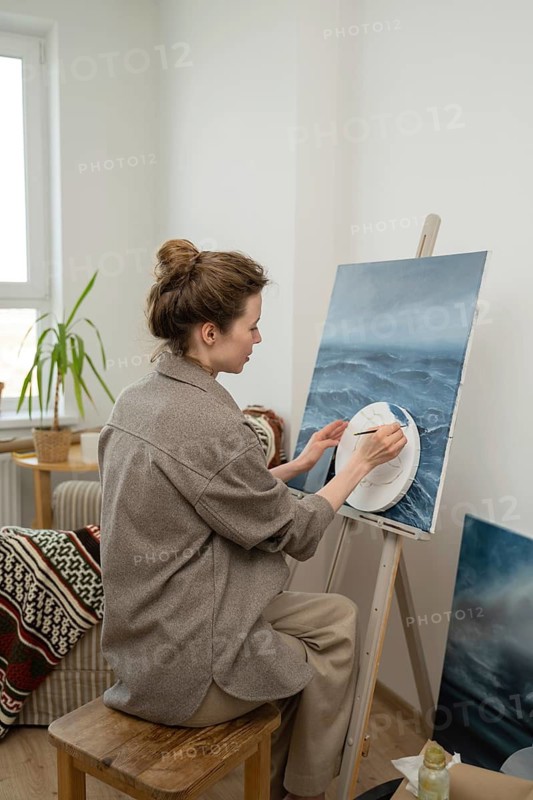 An artist sits on a wooden stool, painting a seascape on a canvas. Surrounded by art supplies, the cozy home studio exudes creativity and inspiration