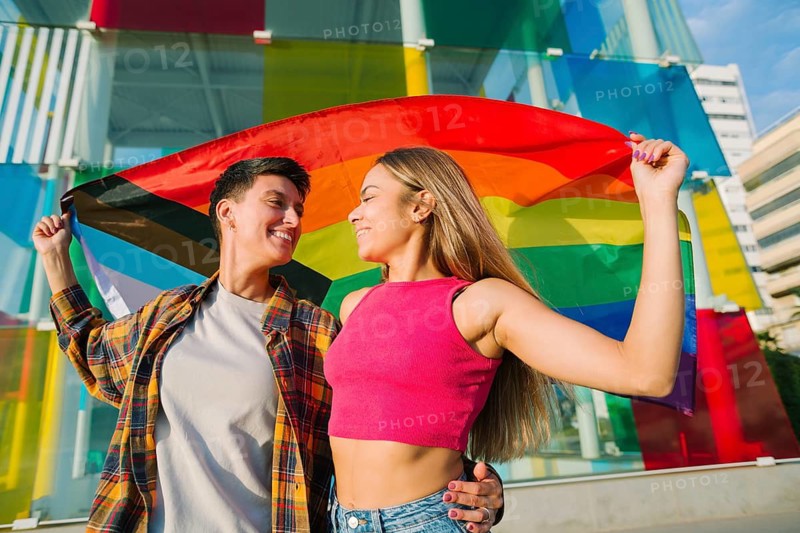 A joyful lesbian couple embraces while holding the Pride and Transgender flags. Their loving gaze and vibrant background symbolize inclusivity, self-expression, and the celebration of diversity