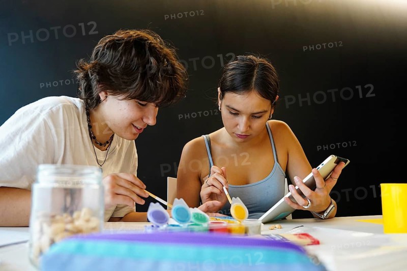 Two teenagers, a non-binary individual and a female, are deeply engaged in a creative art project in a classroom environment. They use various colors for painting, demonstrating a mix of concentration and casual interaction