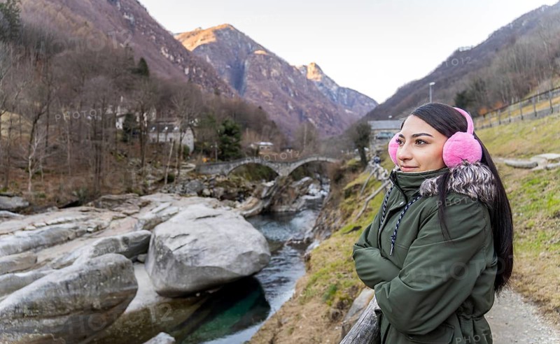 A woman in pink earmuffs and a green jacket admires the picturesque winter scenery of Ponte Dei Salti in Lavertezzo, Ticino, with clear blue water and ancient stone bridge