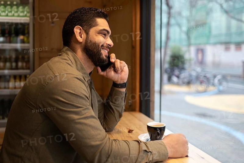 A man in a brown shirt smiles while talking on the phone, sitting at a cafe window with a cup of coffee. Outside, bicycles are visible, creating a relaxed atmosphere in the city