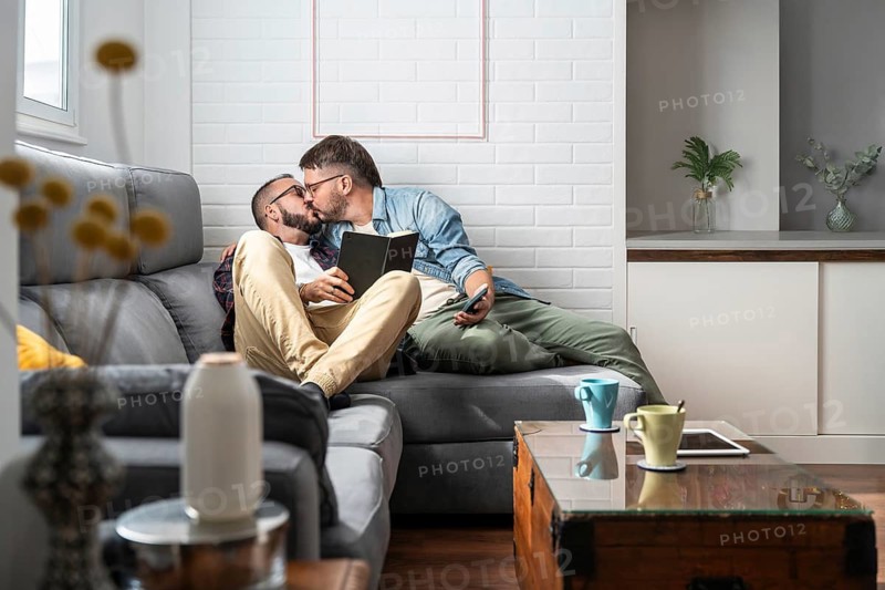 A loving gay couple shares an intimate moment kissing on a comfortable sofa in a modern living room, enjoying each other's company with a book and tablet at hand