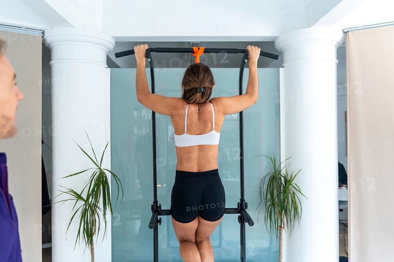 Woman performing a pull up on an exercise bar while a therapist supervises, focusing on rehabilitation, strength training, and medical recovery in a clinic