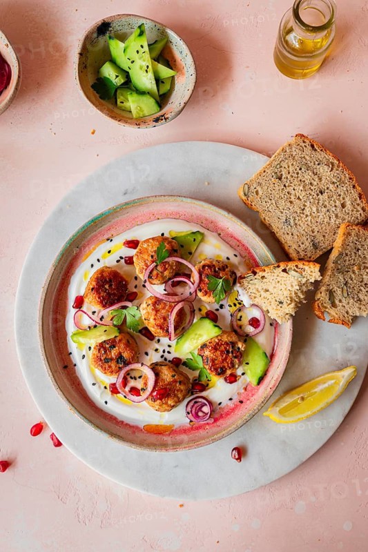 A beautifully presented meatballs dish garnished with cucumber, onion, parsley, and pomegranate on yogurt. Served with crusty bread, cucumber salad, and olive oil