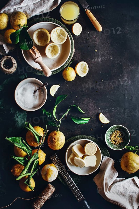 Fresh lemons with zest tool, a bowl of sugar, butter slices, and a glass of juice are arranged on a rustic kitchen table, ready for making delicious lemon curd