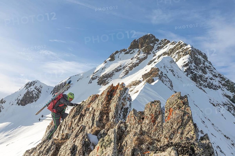 A ski tourer ascends a rugged, snow-covered peak, showcasing determination and adventure against a breathtaking mountainous backdrop under a clear blue sky
