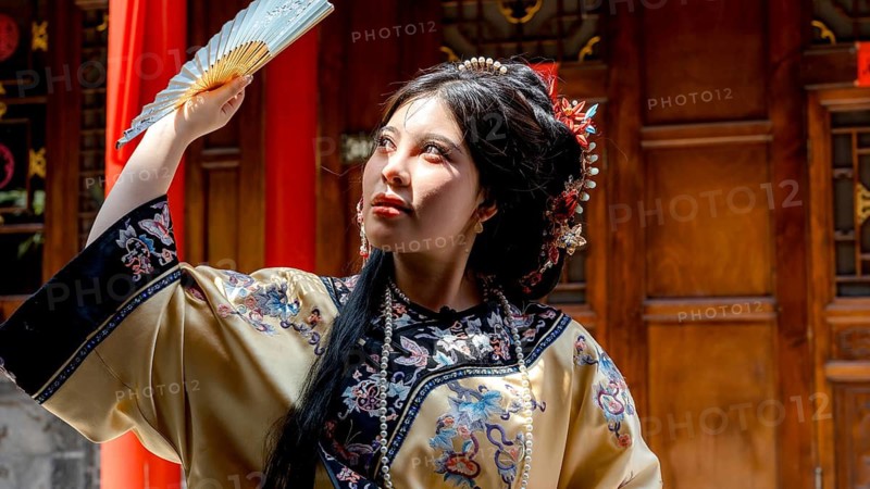 A young woman dressed in traditional Qing Dynasty attire poses with a fan in Pingyao, China. Her elaborate hairstyle and clothing reflect the rich cultural heritage