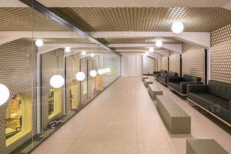 Spacious club hallway featuring a geometric ceiling and mirrored walls with round lights. Minimalist seating and modern interior design create a sleek, stylish atmosphere