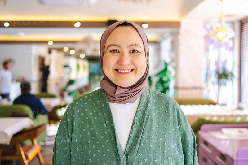 Asian woman wearing a green hijab smiling warmly in a cozy cafe. Her serene expression reflects cultural beauty and traditional attire, emphasizing ethnic diversity