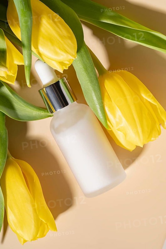A minimalistic composition of a skincare dropper cosmetic surrounded by vibrant yellow tulips on a soft beige surface, evoking a fresh and natural aesthetic