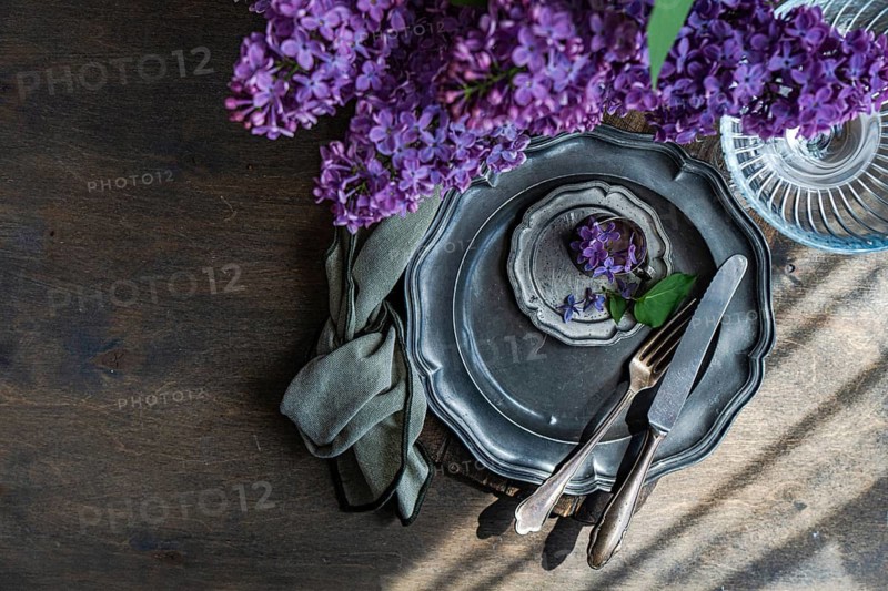 Overhead view of an elegant table setting featuring a vintage metal plate, fork, and knife. A sprig of lilac flowers adds a touch of rustic beauty to the wooden table